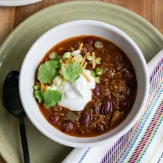 a white bowl filled with chili and sour cream on top of a plate next to a spoon