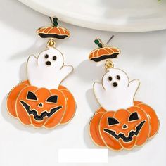 two halloween earrings with pumpkins and ghost faces hanging from the ear wires on a white surface