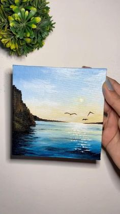 a person holding up a small painting with seagulls flying over the water at sunset