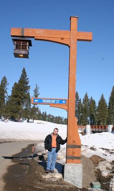 a man standing under a wooden cross on the side of a snow covered road with trees in the background