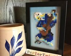 a cross stitch picture next to a potted plant on a wooden table with a blue and white vase