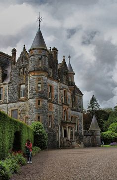 an old castle like building surrounded by greenery and trees with two people walking in front