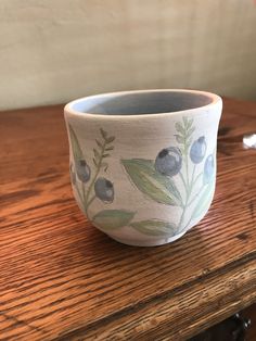 a ceramic bowl with blue berries and leaves painted on the side sitting on a wooden table