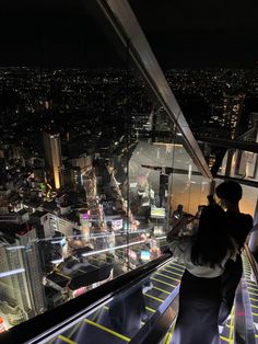 two people standing on top of a tall building looking down at the city lights and skyscrapers