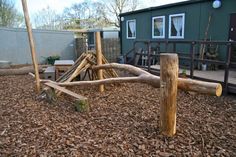 an outdoor play area with wood logs and climbing frame in the foreground, surrounded by mulch