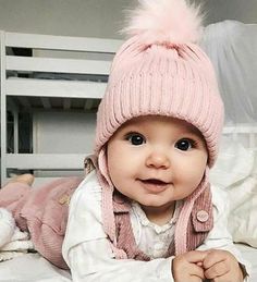 a baby laying on top of a bed wearing a pink hat and jacket with pom - poms