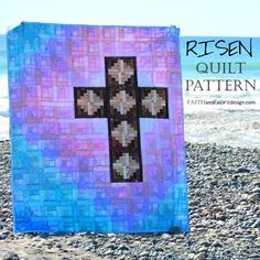 a blue and purple quilt with a cross on it, sitting on some rocks near the water