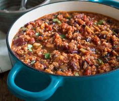 a blue pot filled with chili and beans on top of a wooden table next to a spoon