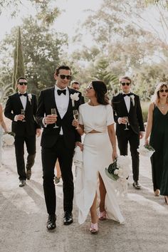 two people in tuxedos are walking with one another and the other is holding champagne glasses