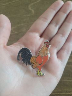 a hand holding a small pin with a rooster on it's chest and tail