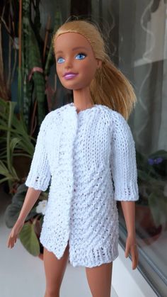 a doll with blonde hair wearing a white knitted sweater and short shorts, standing in front of a window