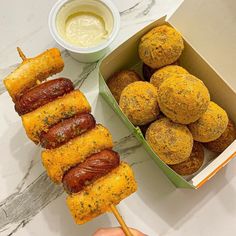 a person holding a hot dog on a stick next to some corn dogs and dipping sauce