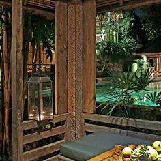 an outdoor table with fruit on it in front of a pool and wooden structure that is surrounded by greenery