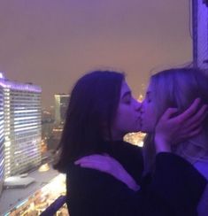 two women kissing each other in front of a cityscape with buildings and lights