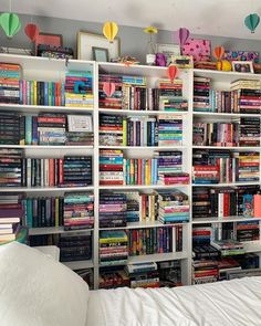 a book shelf filled with lots of books on top of a white bed covered in pillows