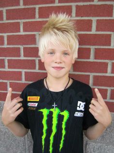 a young boy with blonde hair wearing a black shirt and holding his hands up in front of him