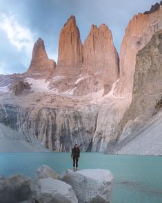 a man standing on top of a large rock next to a lake and mountain range