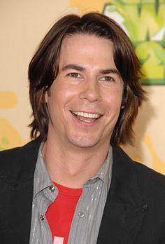 a man with long hair wearing a black jacket and red shirt smiles at the camera