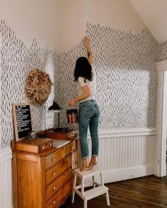 a woman standing on top of a stool in front of a wooden dresser and wallpaper