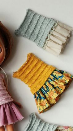 the doll is next to four different knitted clothes for her dolls, which are also handmade