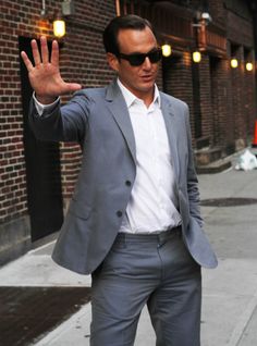 a man in a gray suit and sunglasses waves to the camera while standing on a city street