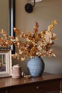 a vase filled with leaves sitting on top of a wooden table next to a mirror