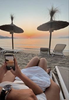 a woman laying on top of a beach next to the ocean holding a cell phone