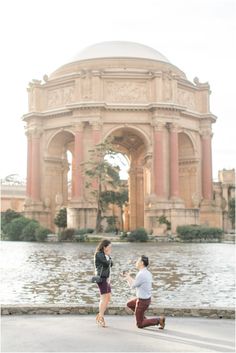 a man kneeling down next to a woman in front of a building with a fountain