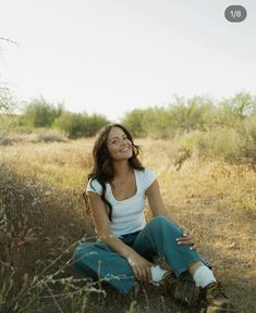 a woman sitting on the ground in a field with tall grass and bushes behind her