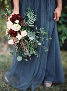 a woman in a long blue dress holding a bouquet of red and white flowers with greenery