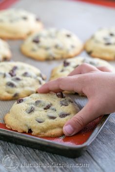 a person picking up a chocolate chip cookie from a baking sheet on a wooden table