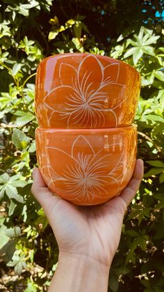 two orange bowls with white flowers on them in front of some green bushes and trees