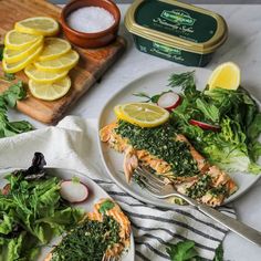 two white plates topped with salmon and greens next to lemon wedges on a cutting board