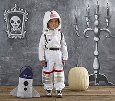 a little boy dressed in an astronaut suit standing next to a chalkboard wall with halloween decorations on it