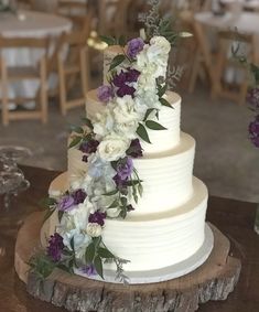 a white wedding cake with purple and white flowers on the top is surrounded by greenery