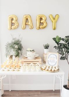 a baby shower party with gold balloons and desserts on a white table next to a potted plant