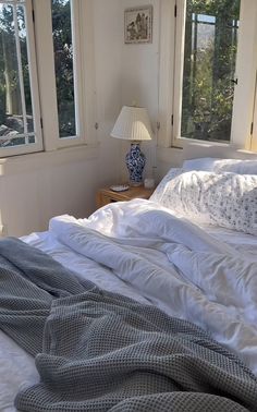 an unmade bed in a bedroom with three windows and a lamp on the nightstand