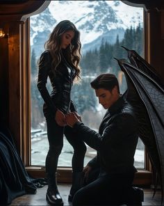 a man and woman dressed in black standing next to each other near a window with snow covered mountains behind them