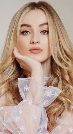 a woman with long blonde hair wearing a white dress and posing for the camera, her hand on her chin