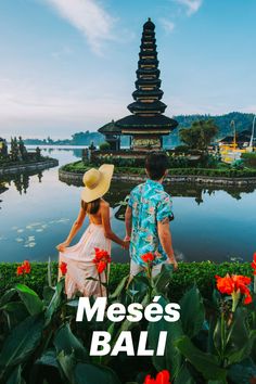 a man and woman standing next to each other in front of water with red flowers