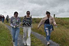 three women walking down a dirt road with grass on both sides and people in the background