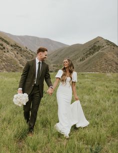 a bride and groom holding hands walking through the grass with mountains in the back ground