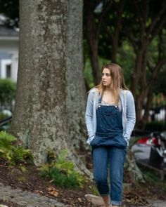 a woman standing in front of a tree wearing overalls and a blue cardigan