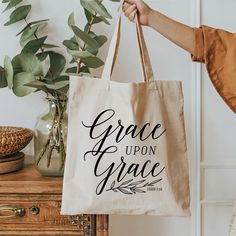 a woman holding a bag with the words grace upon grace printed on it, next to a potted plant