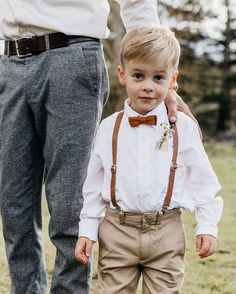 a young boy wearing a bow tie and suspenders standing next to an adult in a field