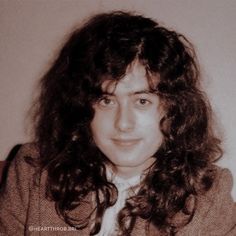 an old photo of a woman with long curly hair and a sweater on, sitting in front of a white wall