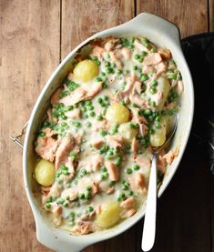 a casserole dish with meat, potatoes and peas on a wooden table next to a spoon