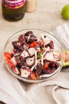 an octopus salad in a glass bowl next to a bottle of ketchup and limes