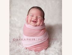 a baby wrapped in a pink wrap smiles while laying on a white blanket with her hands under her chin