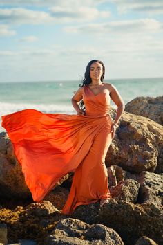 a woman in an orange dress standing on rocks near the ocean